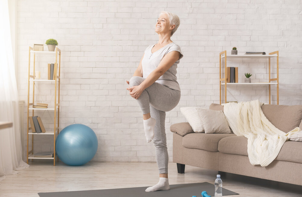 Elderly woman stretching in living room with yoga ball nearby.